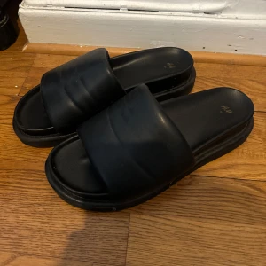 Svarta sandaler från H&M - Säljer ett par svarta sandaler från H&M i mycket bra skick. De har en bred rem över foten och en bekväm sula, perfekta för sommardagar. Passar både till stranden och en avslappnad dag i stan. 