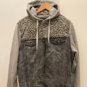 Säljer en unik trendig stiliga grå jeansjacka med huva och leopardmönstrad överdel. Jackan har gråa ärmar och en cool kombination av denim och mjukt tyg. Perfekt för höst och vår, med knappar framtill och två bröstfickor. Passar både till vardags och festliga tillfällen