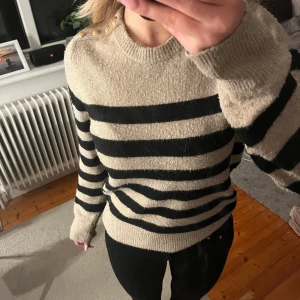 Randig tröja med knappar på axeln - Säljer en superfin randig tröja i beige och svart. Den har långa ärmar och snygga knappar på axeln som ger en extra touch. Perfekt för höst och vinter, och den är i mycket bra skick. Passar både till vardags och lite mer uppklädda tillfällen.