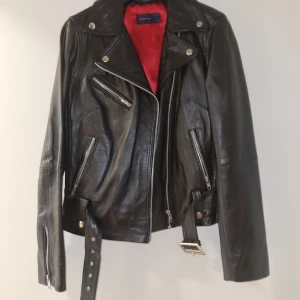 Svart skinnjacka från Rock'n Blue - Säljer en snygg svart skinnjacka från Rock'n Blue i storlek 36. Jackan har en cool bikerstil med dragkedjor och bälte i midjan. Insidan är fodrad med rött tyg som ger en härlig kontrast. Perfekt för höst och vår! Oanvänd! 
