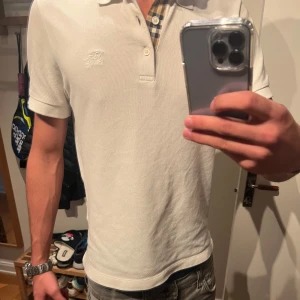 Vit pikétröja från Burberry - Säljer en stilren vit pikétröja från Burberry i storlek M. Tröjan har klassisk krage med knappar och ett diskret broderat märke på bröstet. Insidan av kragen har det ikoniska Burberry-mönstret. Perfekt för en elegant och avslappnad look.