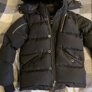 Moose Knuckles - Säljer en svart dunjacka från Moose Knuckles i mycket bra skick. Jackan har en pufferstil med dragkedja och knappar, samt coola detaljer som metalliska dekorationer på ärmarna. Perfekt för kalla vinterdagar! 🖤