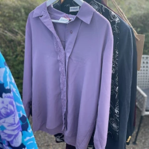 Lila blus från Spaderdam - Säljer en elegant lila blus från Spaderdam i storlek 42. Blusen har långa ärmar och en fin spetsdetalj längs knäppningen. Perfekt för både vardag och festliga tillfällen. 💜