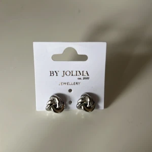 Silverfärgade örhängen från By Jolima - Helt nya! Aldrig använda! Snygga och stilrena silverfärgade örhängen från By Jolima. De har en elegant knutdesign som passar perfekt för både vardag och fest. Perfekta för att ge din outfit en extra touch av elegans. Köp dem nu och lyft din stil! ✨