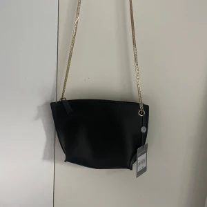 Svart axelväska med guldkedja - Snygg svart axelväska i syntetmaterial med guldfärgad kedja som axelrem. Väskan har en dragkedja upptill och är i nytt skick med etikett kvar. Perfekt för både vardag och fest! I nyskick med prislapp kvar!