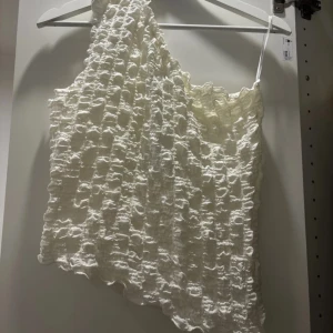 Vit oneshoulder topp - Säljer en superfin vit oneshoulder topp med ett unikt texturerat mönster. Perfekt för sommarens fester eller en dag på stan. Toppen är i ett lätt och luftigt material som gör den bekväm att bära hela dagen. Passar perfekt till både jeans och kjol! Prislapp på!