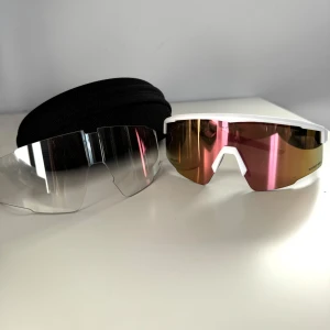 Sportiga solglasögon från Everest  - Säljer ett par sportiga solglasögon med vit båge och spegelglas i rosa och guld. Perfekta för träning och utomhusaktiviteter. Kommer med extra klara linser och ett praktiskt fodral. Glasögonen har en modern och aerodynamisk design.