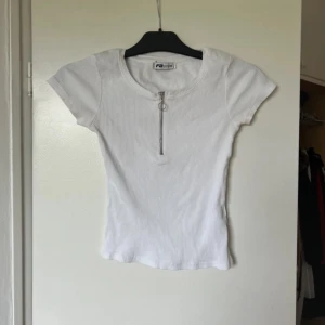 Vit ribbad topp - Säljer en superfin vit ribbad topp. Den har en dragkedja framtill och korta ärmar, perfekt för sommardagar. Toppen är i mycket bra skick och passar både till vardags och fest. Skön och stilren design som är lätt att matcha med andra plagg.