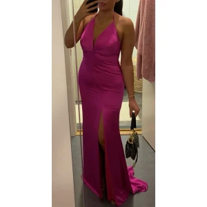 Rosa satin långklänning med slits - Säljer en fantastisk rosa långklänning i satin med en elegant slits framtill. Klänningen har en djup v-ringning och är ärmlös, perfekt för festliga tillfällen. Den sitter tight och framhäver figuren på ett smickrande sätt. Perfekt för bröllop och förlovning eller andra speciella tillfällen!