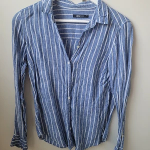 Blå skjorta från Gina Tricot - Säljer en snygg blå- och vitrandig skjorta från Gina Tricot i storlek 36. Skjortan har långa ärmar och knappar framtill. Perfekt för både vardag och festliga tillfällen. Den är i mycket bra skick och gjord av ett skönt material som känns lätt mot huden.