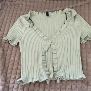 Ljusgrön ribbad kofta från H&M Divided - Säljer en superfin ljusgrön kofta från H&M Divided. Den är ribbad med volangdetaljer och kortärmad, perfekt för vår och sommar. Koftan har en knapp framtill och är i mycket bra skick. Passar perfekt till både vardag och fest! 🌿