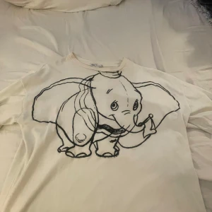 Vit t-shirt med elefantmotiv från Zara - Säljer en söt vit t-shirt från Zara med ett stort elefantmotiv på framsidan. T-shirten är i samarbete med Disney och har korta ärmar. Perfekt för en avslappnad stil eller som en rolig detalj i vardagsoutfiten. 🐘