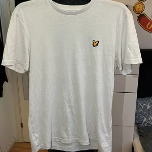 Vit Lyle & Scott t-shirt - Säljer en stilren vit t-shirt med en liten gul logga på bröstet. Perfekt för vardagsbruk eller som en basplagg i garderoben. T-shirten har en normal passform och är kortärmad, vilket gör den idealisk för varmare dagar.