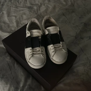 Valentino sneakers i vitt och svart - Säljer ett par snygga Valentino sneakers i vitt med en bred svart rem över. De är i bra skick med lite slitage på sulan. Perfekta för en stilren look och passar till både vardag och fest. Skorna har snörning och är gjorda i skinn. Du får även me lådan och en påse som äktighetsbevis. Priset går att diskutera!