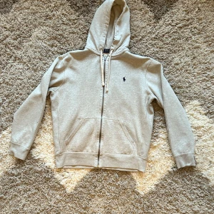 Grå zip-up hoodie, Polo Ralph Lauren - Säljer en snygg Grå zip-up hoodie från Polo Ralph Lauren i storlek M. Den har en dragkedja framtill och en klassisk broderad logga på bröstet. Perfekt för höst och vår, gjord i mjuk bomullsmix för extra komfort. 