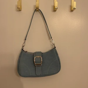 Blå axelväska med spänne - Snygg blå axelväska i jeansmaterial med ett stort dekorativt spänne på framsidan. Väskan har en dragkedja upptill och en justerbar axelrem. Perfekt för vardagsbruk eller en dag på stan!