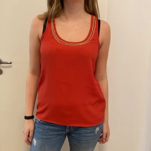 Röd ärmlös topp från Cotton On - Snygg röd ärmlös topp från Cotton On i storlek S. Den har en cool kedjedetalj runt halsringningen som ger en extra touch. Perfekt för sommardagar eller en avslappnad utekväll. Toppen är i bomull och känns skön mot huden.
