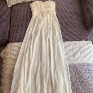 Vit axelbandslös långklänning - Elegant vit axelbandslös långklänning perfekt för sommarens fester eller bröllop. Klänningen har en vacker drapering och en smickrande passform. Materialet är mjukt och faller fint, vilket ger en romantisk känsla. Perfekt för en speciell dag!