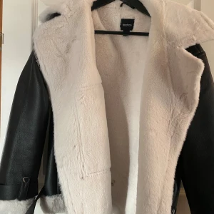 Svart jacka med vit fuskpäls från Boohoo - Säljer en snygg svart jacka från Boohoo med vit fuskpäls på insidan. Jackan har långa ärmar och dragkedja. Perfekt för höst och vinter, håller dig varm och stilren. Passar bra till både vardag och fest!