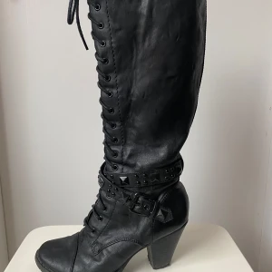 Svarta höga boots med snörning och nitar - Säljer ett par svarta höga boots  med snörning och coola nitar runt ankeln. De har en rejäl klack och dragkedja på sidan för enkel på- och avtagning. Perfekta för höst och vinter när du vill ha en edgy look. Passar både till jeans och klänning!