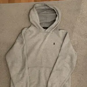 Säljer en stilren grå hoodie från Polo Ralph Lauren. Den har en klassisk design med en liten broderad logga på bröstet. Perfekt för både vardag och chill. Den är i bra skick och superbekväm att ha på sig. Passar perfekt till jeans eller joggers!