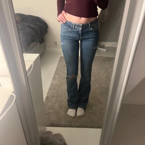 Blå jeans med slitningar - Säljer ett par snygga blå jeans med slitningar på knäna. De är lågmidjade och har en bootcut-stil som ger en avslappnad look. Perfekta för en casual outfit!