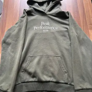 Säljer en snygg olivgrön hoodie från Peak Performance. Den har en klassisk design med en känguruficka framtill och en broderad logga på bröstet. Perfekt för både höst och vår, och den är superbekväm att ha på sig. Passar perfekt för en avslappnad stil!