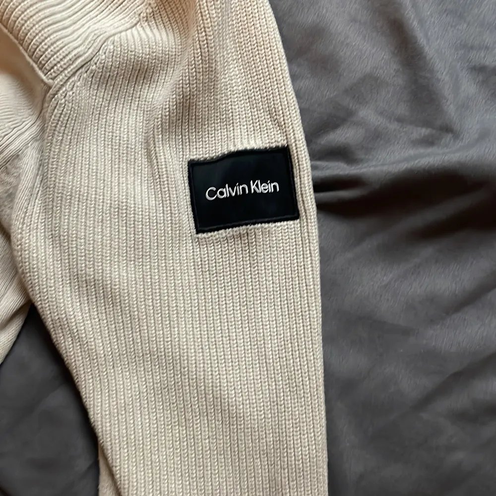 Säljer en snygg beige tröja från Calvin Klein. Tröjan är ribbad på ärmarna och har en stilren design med en liten Calvin Klein-logga på ärmen. Perfekt för både vardag och lite finare tillfällen! Litet hål under högra armen . Tröjor & Koftor.