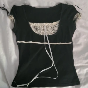 Svart topp med spetsdetaljer - Säljer en svart topp med vackra spetsdetaljer vid halsringningen. Toppen har korta ärmar och en söt knytning framtill. Perfekt för en somrig look eller en festlig kväll!