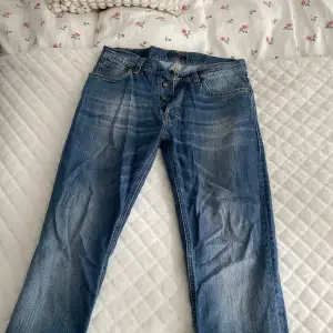 Säljer ett par snygga blå jeans med knappar framtill. De har en klassisk femficksdesign och en lätt slitning för en cool look. Perfekta för vardagsbruk!