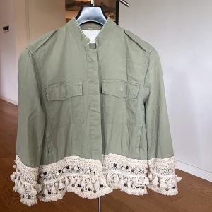 Olivgrön jacka med spetsdetaljer från Zara Woman - Säljer en superfin olivgrön jacka från Zara Woman. Den har unika spetsdetaljer och fransar längst ner som ger en bohemisk look. Jackan är bra för höst och vår, normal passform. Den är tillverkad i ett mjukt bomullsmaterial . Två fickor