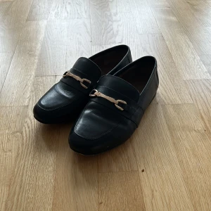 Svarta loafers med metallspänne - Säljer ett par stilrena svarta loafers i skinn med ett snyggt metallspänne framtill. Perfekta för både vardag och fest. De har en klassisk design och är superbekväma att ha på sig. Passar till nästan alla outfits!