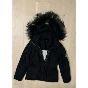 Svart parkajacka med pälsdetaljer - Säljer en snygg svart Canadiens dunjacka med pälsdetaljer på huvan. Jackan har en dragkedja och knappar framtill, vilket gör den perfekt för kalla vinterdagar. 