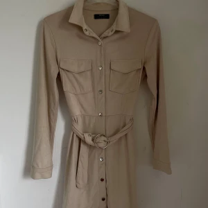 Beige klänning från Bershka - Säljer en superfin beige jacka från Bershka i storlek XS. Den har långa ärmar och en snygg krage. Den har knappar hela vägen ner och ett bälte i midjan som ger en fin passform. Perfekt för vardag.
