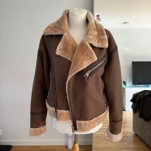 Brun pälsjacka med dragkedja - Säljer en supermysig brun pälsjacka med fluffigt foder och coola dragkedjedetaljer. Jackan har en hög krage som kan stängas med en rem och är perfekt för kalla dagar. Den är långärmad och har en snygg, lite oversized passform. Perfekt för höst och vinter!