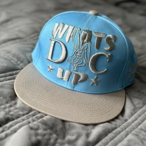 Blå keps från State Property - Keps från State Property med texten 'What's Up Doc' och en tecknad kanin broderad på framsidan. Kepsen har en grå skärm och justerbar baksida.