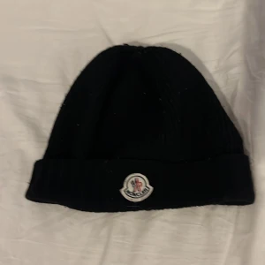 Svart mössa från Moncler - Säljer en snygg svart mössa från Moncler. Den är stickad och har en klassisk design med Monclers logga framtill. Perfekt för att hålla sig varm under kalla dagar!