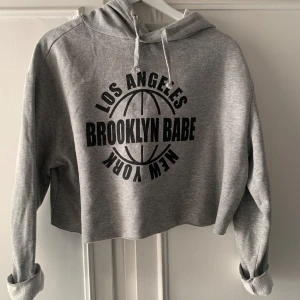 Grå croppad hoodie med tryck - Säljer en supercool grå croppad hoodie med text i svart. Perfekt för en avslappnad stil och passar till nästan allt. Den har långa ärmar och en skön huva med snören. 