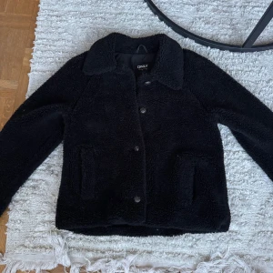 Svart teddyjacka från ONLY - Säljer en supermysig svart teddyjacka från ONLY i storlek M. Jackan har en klassisk krage och knappar framtill. Perfekt för kyliga höst- och vinterdagar 