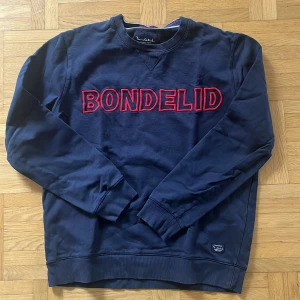Mörkblå sweatshirt från Bondelid - Säljer en snygg mörkblå sweatshirt från Bondelid med röd broderad text på framsidan. Tröjan är i storlek M och har långa ärmar. Perfekt för en casual look och passar bra till både jeans och chinos.