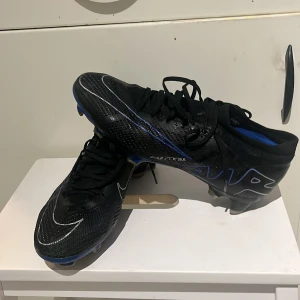 Nike fotbollsskor  - Jag säljer ett par Nike  AIR ZOOM MERCURIAL VAPOR 15 PRO FG SHADOW - SVART/KROM/BLÅ stl 41 26cm. Jag har andvänt skora 1 träning och jag säljer skorna för det vart för små innan fotbollssäsongen började. Orginal pris 1800kr 