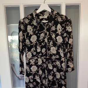 Svart blommig klänning från Zara Woman - Säljer en superfin svart klänning från Zara Woman med ett vackert blommigt mönster i vitt. Klänningen har långa ärmar och en klassisk krage, perfekt för både vardag och fest.