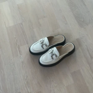 Vita loafers från K.Cobler - Säljer ett par stilrena vita loafers från K.Cobler. De har en snygg silverdetalj framtill och en svart sula som ger en cool kontrast. Perfekta för både vardag och fest!