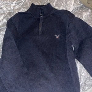 Mörkblå stickad tröja från GANT - Säljer en snygg och stilren mörkblå stickad tröja från GANT. Den har en praktisk dragkedja vid halsen och är perfekt för både höst och vinter. Tröjan är i storlek M och är i bra skick. Perfekt för att hålla sig varm och se bra ut samtidigt!