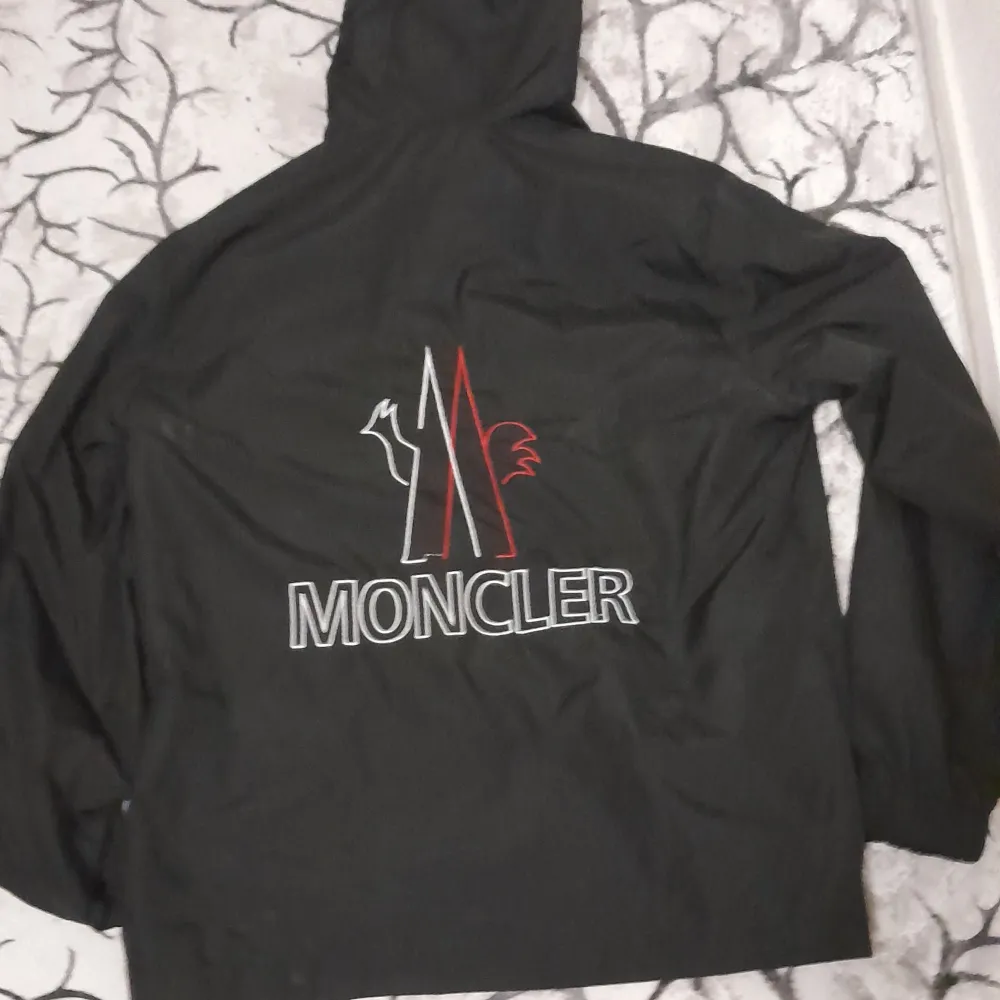 Säljer en snygg svart vindjacka från Moncler. Jackan har en dragkedja framtill och en huva med textdetaljer. Den har också Monclers logga på bröstet och en stor logga på ryggen. Perfekt för blåsiga dagar och ger en stilren look. Passar för L och M. Jackor.