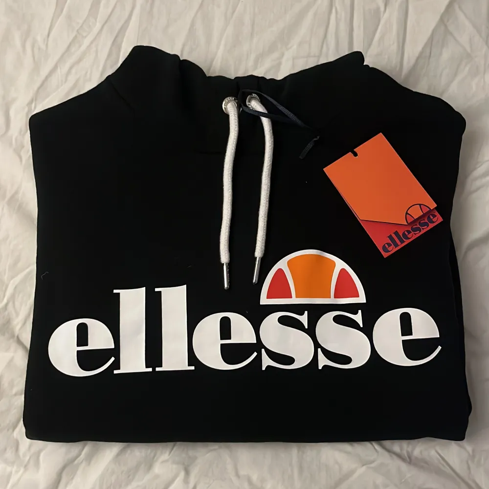 Säljer en snygg och bekväm svart hoodie från Ellesse. Den har en stor logga på framsidan i vitt och orange, och en praktisk känguruficka. Perfekt för en avslappnad stil eller för att hålla sig varm under kyliga dagar. Huvan har vita dragsnören för justering. Helt ny med tag kvar!. Hoodies.