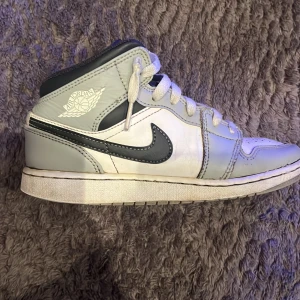 Nike Air Jordan 1 Mid (GS) i grå och vit - Säljer ett par Nike Air Jordan 1 Mid (GS) i färgerna ljusgrå och vit. De är i bra skick. Men det enda felet är ett inuti vid hälen är det slitet/ett litet hål in i tyget. Inte creased har andvänt creas skydd har impregnering. Men annars väldigt fina😍
