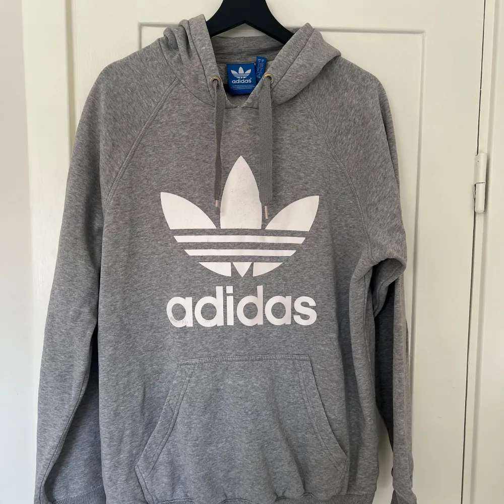 Säljer en klassisk grå hoodie från Adidas med det ikoniska trefoil-loggan i vitt på framsidan. Den är superbekväm och perfekt för både chill och träning. Hoodien har en stor ficka fram och justerbar dragsko i huvan. Perfekt för höst och vinter!. Tröjor & Koftor.