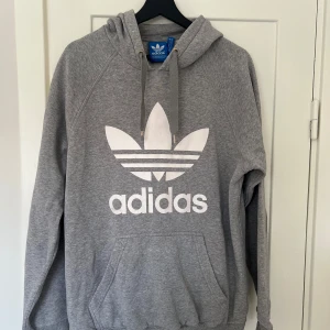 Grå hoodie från Adidas - Säljer en klassisk grå hoodie från Adidas med det ikoniska trefoil-loggan i vitt på framsidan. Den är superbekväm och perfekt för både chill och träning. Hoodien har en stor ficka fram och justerbar dragsko i huvan. Perfekt för höst och vinter!