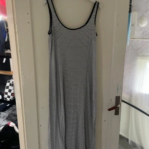 Randig långklänning med svarta detaljer - Säljer en superfin randig långklänning i svart och vitt. Den har en enkel och stilren design med svarta kanter vid ärmhålen och halsringningen. Perfekt för en avslappnad sommarlook eller en dag på stranden. Klänningen är ärmlös och har en bekväm passform.